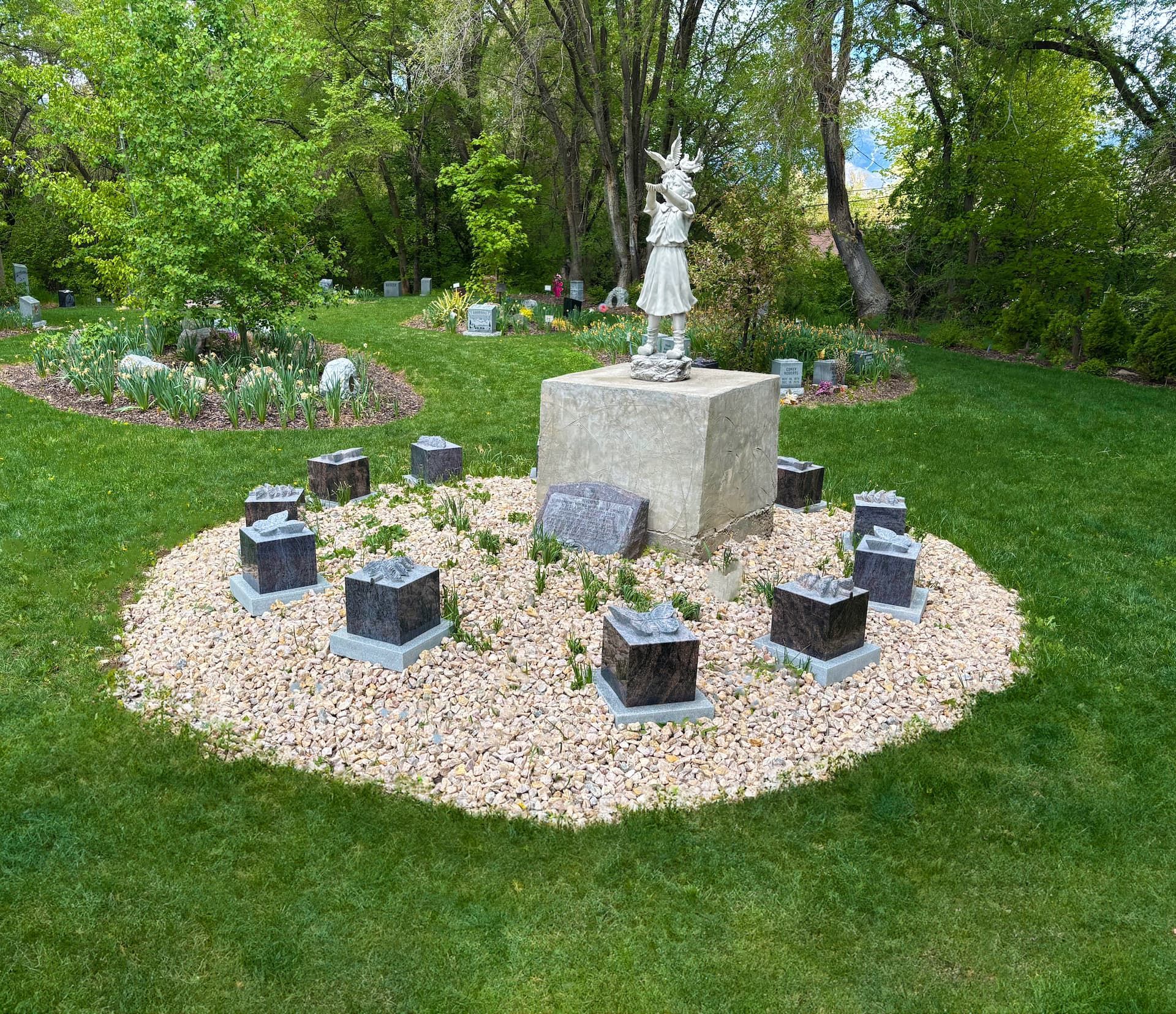 Small outdoor memorial with a central statue on a stone pedestal, ringed by black markers on gravel in a grassy park.