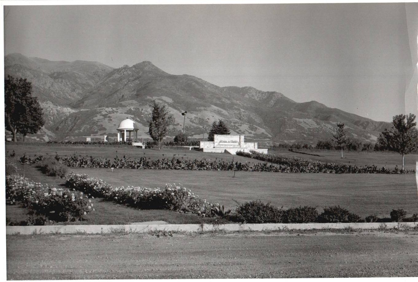 Black-and-white rural landscape with fields, a white building, a water tower, and mountains in the background