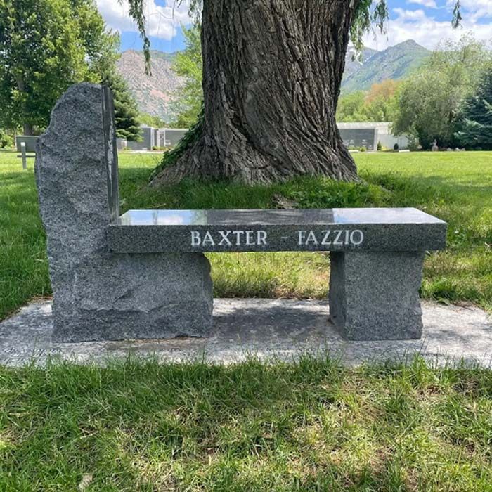 Stone bench engraved “BAXTER - FAZZIO” beside a large tree in a grassy park with mountains in the background