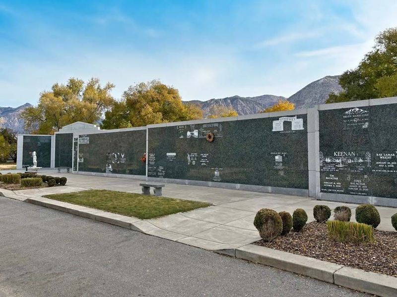 Modern memorial wall with engraved names, sidewalk, trimmed shrubs, and mountains under a blue sky.