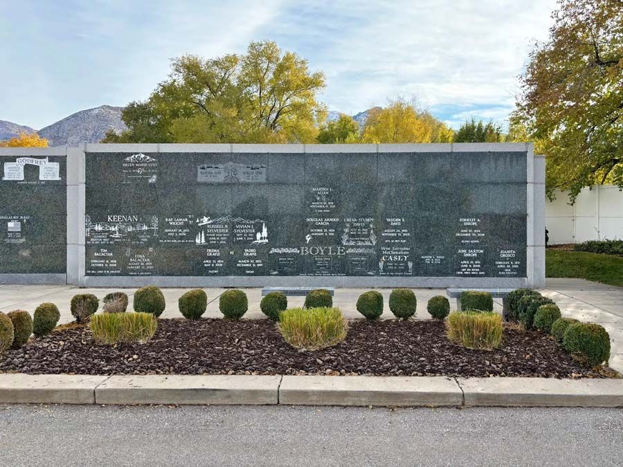 Outdoor memorial wall with engraved names, bordered by shrubs and autumn trees under a cloudy sky