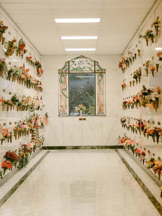 White memorial wall lined with flowers and photos, framing a central window in a bright hallway