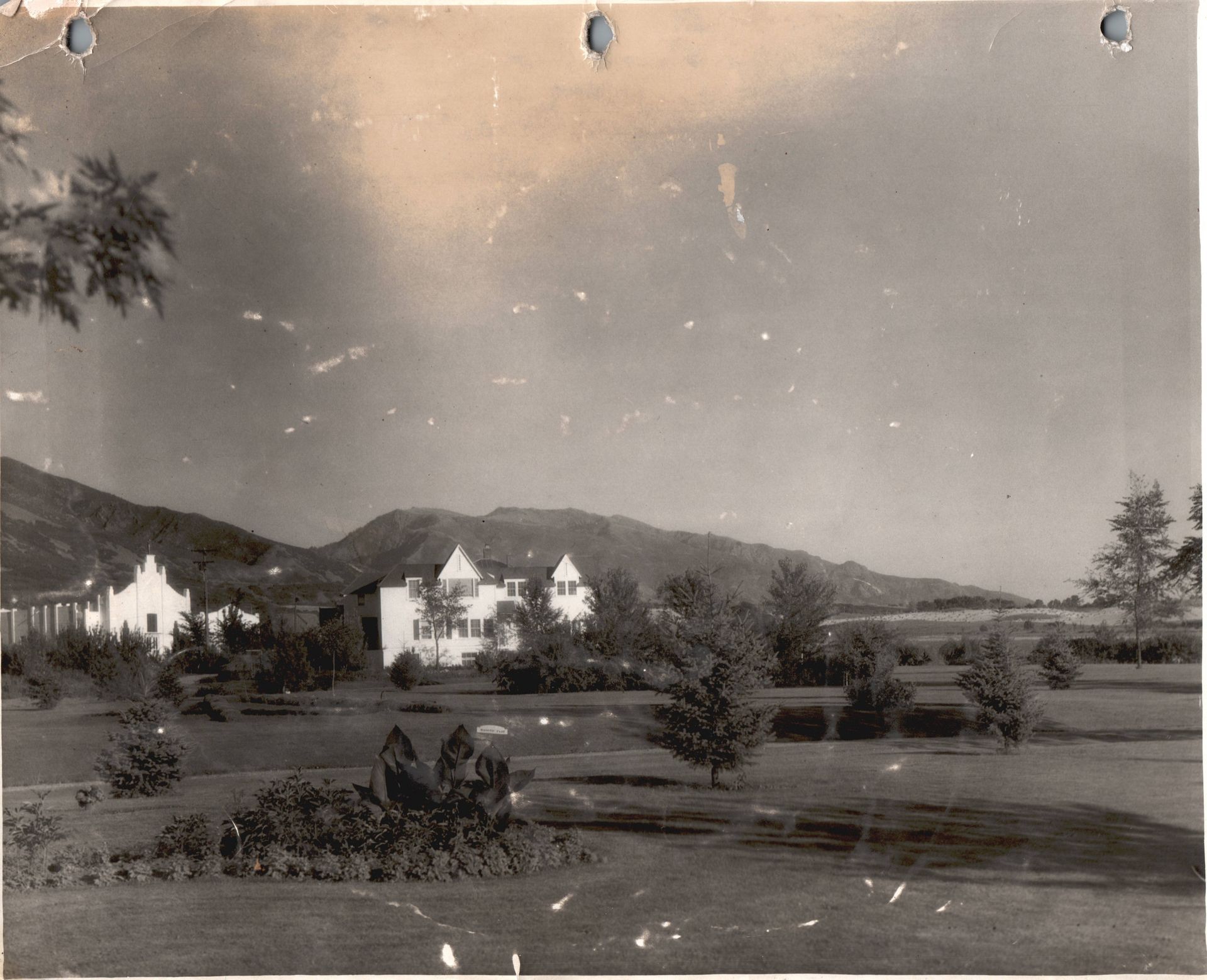 Vintage black-and-white photo of a large house in a rural landscape with mountains and trees in the background