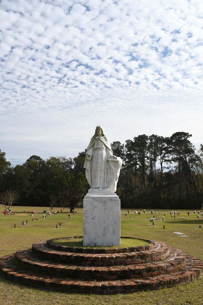 White statue on a brick pedestal in a grassy cemetery under a cloudy sky