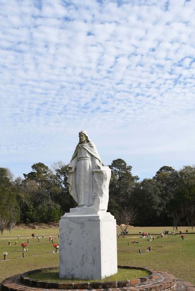 White statue on a pedestal in a grassy cemetery, with trees and a cloudy sky in the background.
