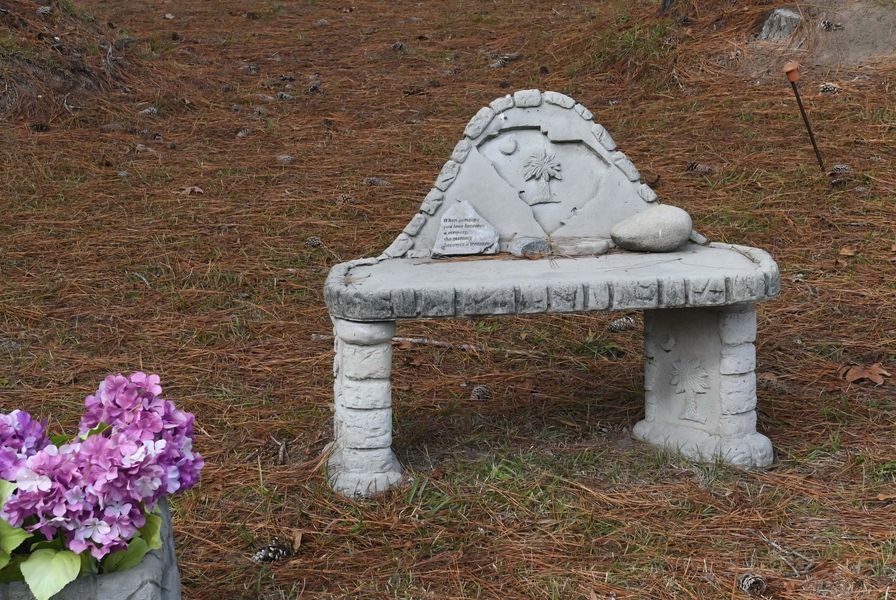 Small white stone bench or memorial in a grassy area, with purple flowers in the lower left.