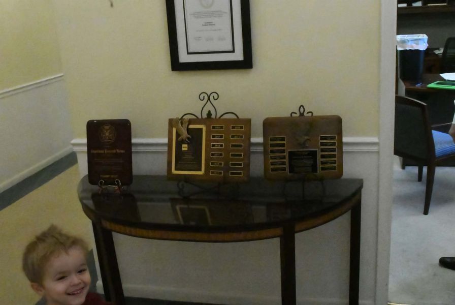 Table with display boxes under a framed picture in a hallway; smiling child peeks in from lower left.