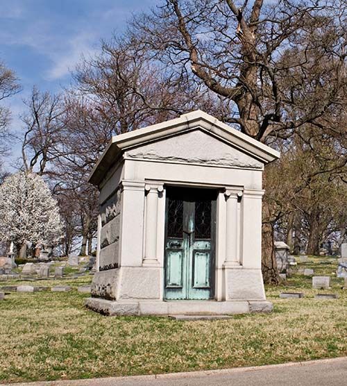 Small stone mausoleum with pale green doors in a cemetery under leafless trees.