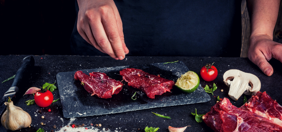 Hands seasoning raw steaks on a dark slate board with garlic, lime, tomato, and mushrooms