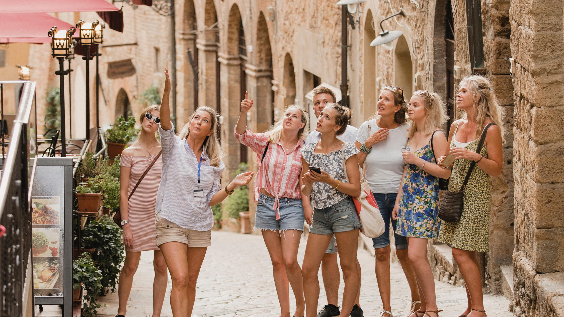 Friends in summer clothes walking and chatting on a sunlit cobblestone street lined with historic buildings.