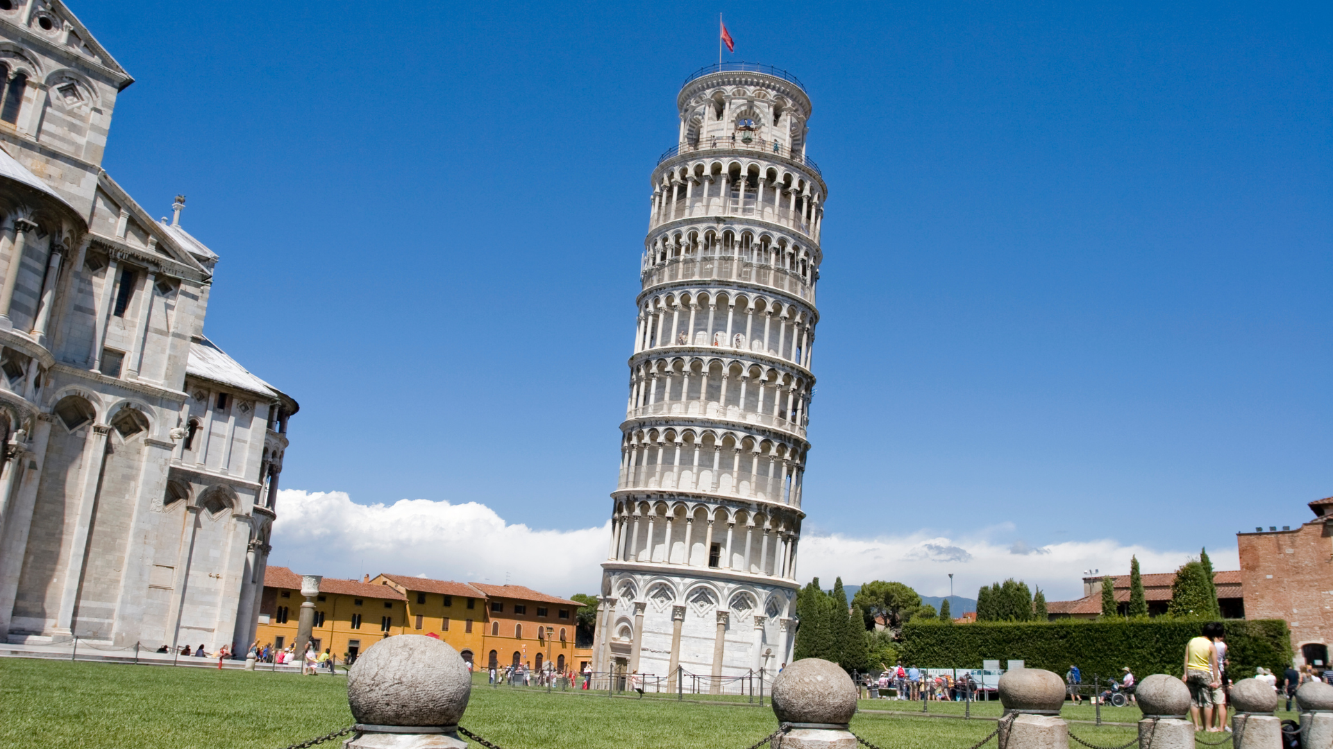Leaning Tower of Pisa in a sunny plaza with green lawn and historic buildings under a blue sky