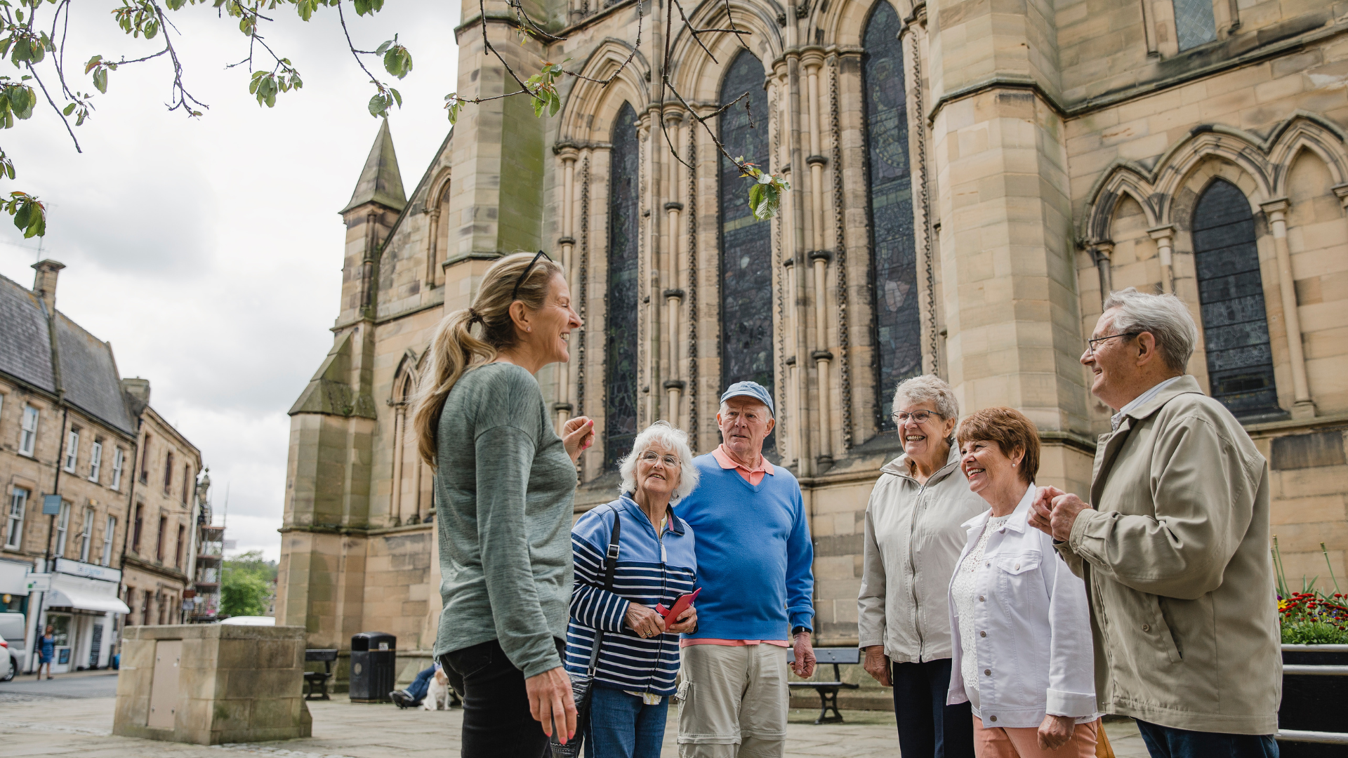 Group touring a stone cathedral with a guide in a courtyard, trees overhead.