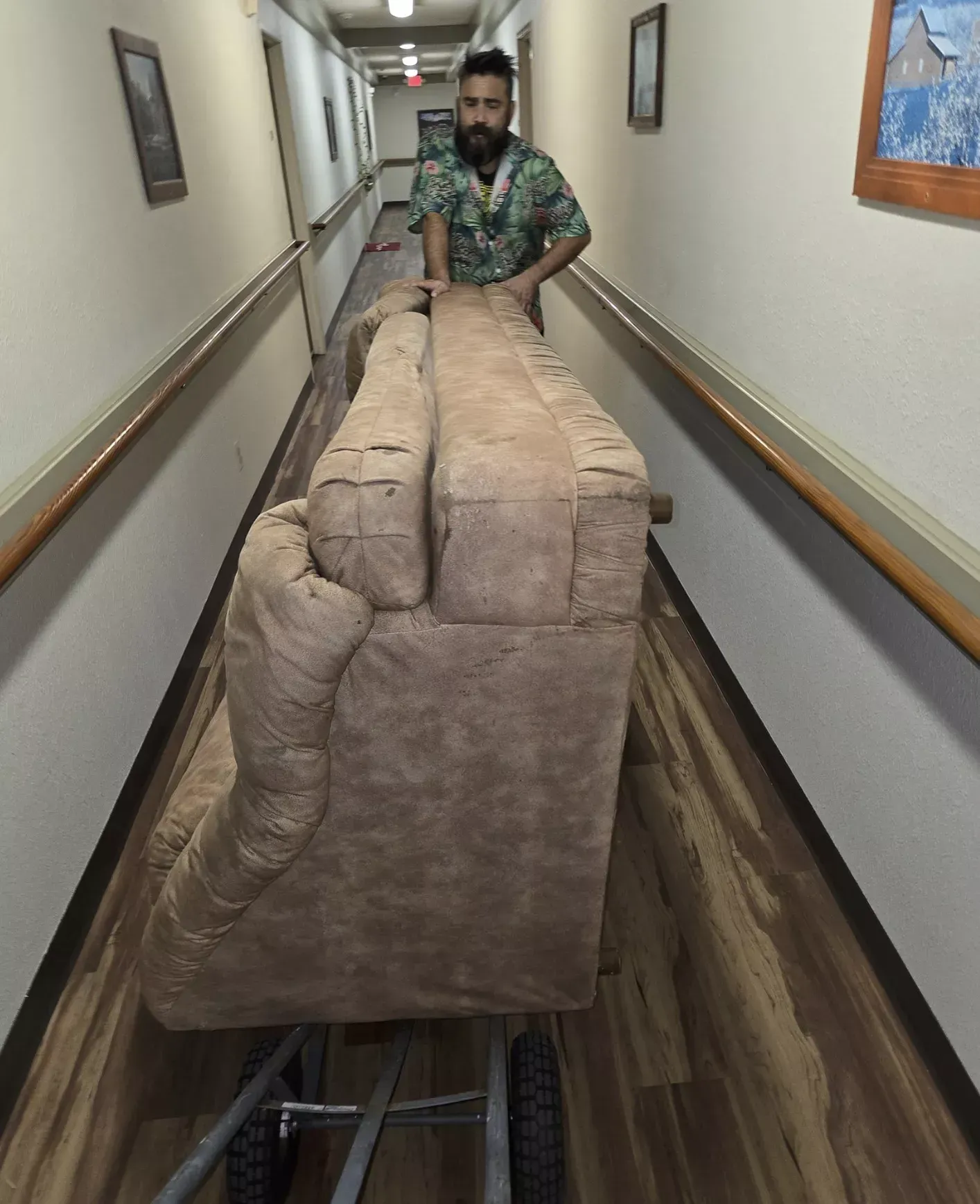 A person pushes a large, tan sofa upright on a hand truck through a long, narrow hallway with wooden flooring.