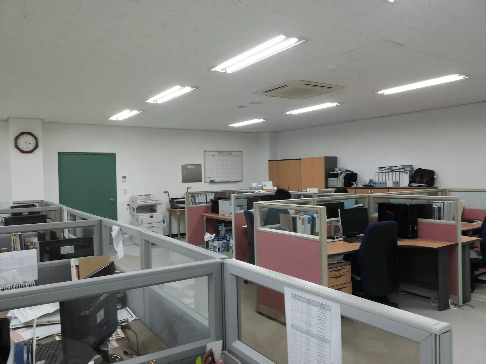 An office space with multiple cubicles, wooden cabinets, and ceiling lights, view taken from the entrance area.