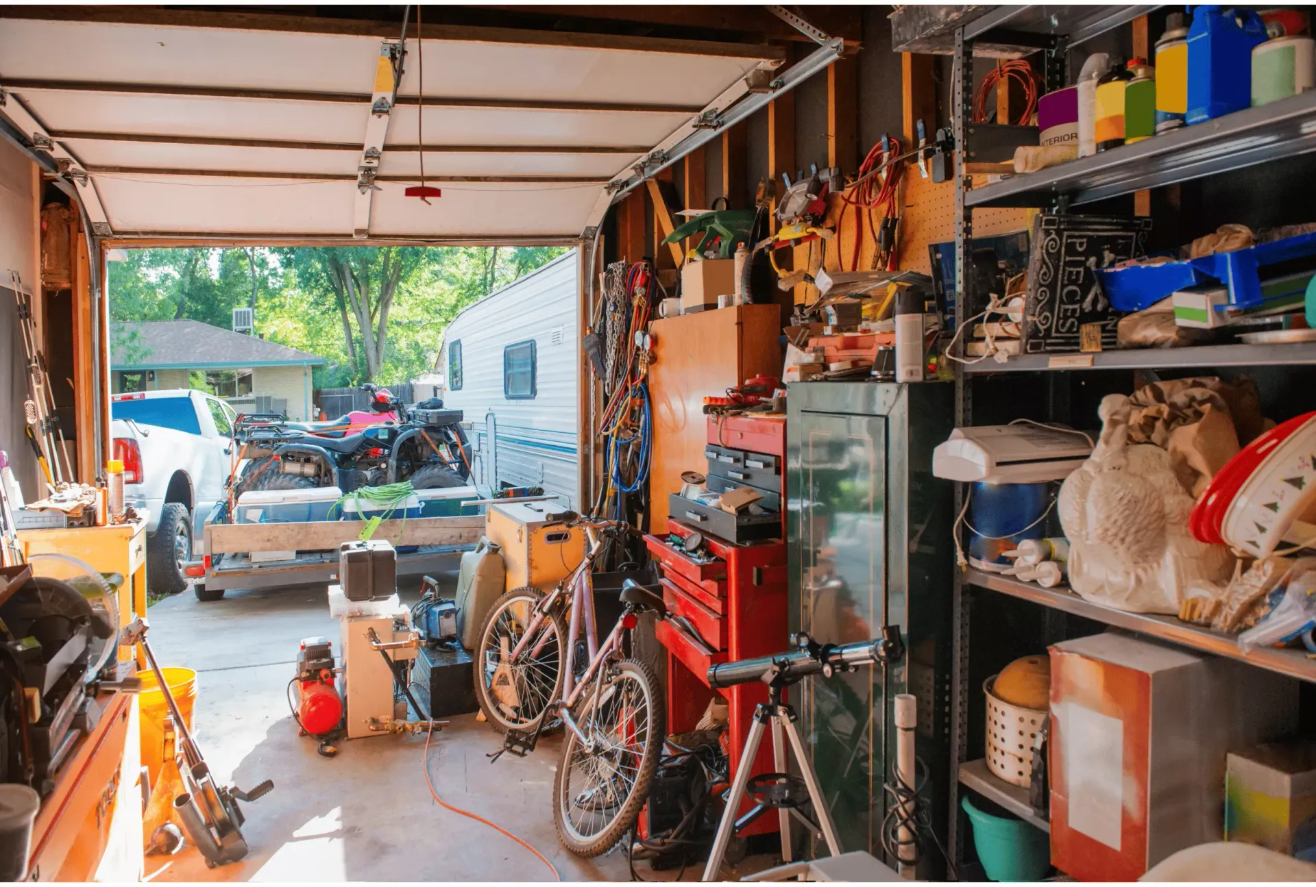 A cluttered garage with shelves of supplies, a bicycle, tools, and an open door revealing a driveway with a truck and RV.