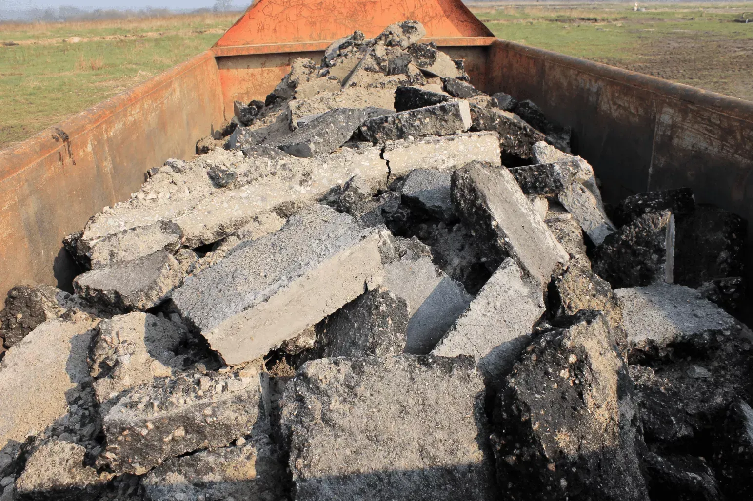 A dump truck bed filled with large chunks of broken asphalt and concrete debris outdoors.