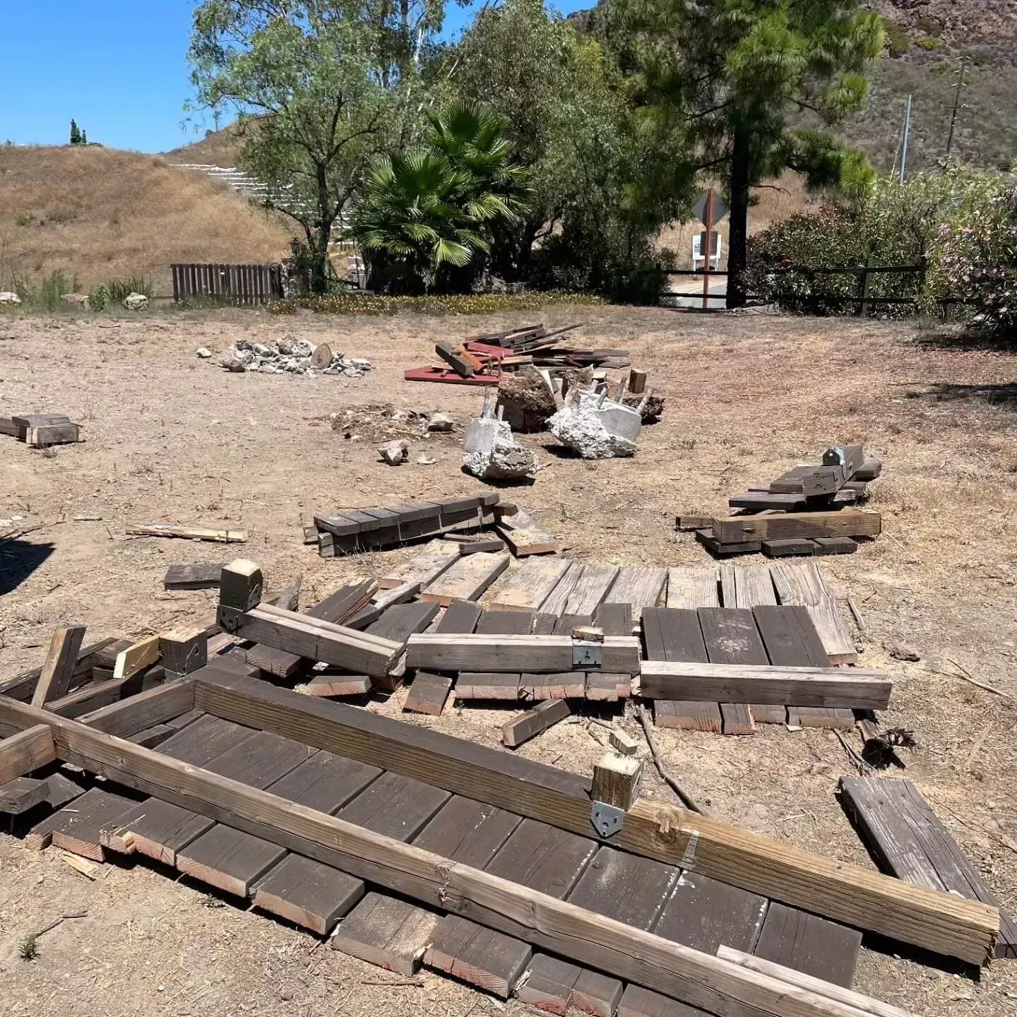 Scattered wood panels, lumber, and debris from a dismantled wooden structure lie on a dry, dirt lot in a rural setting near Ventura, CA
