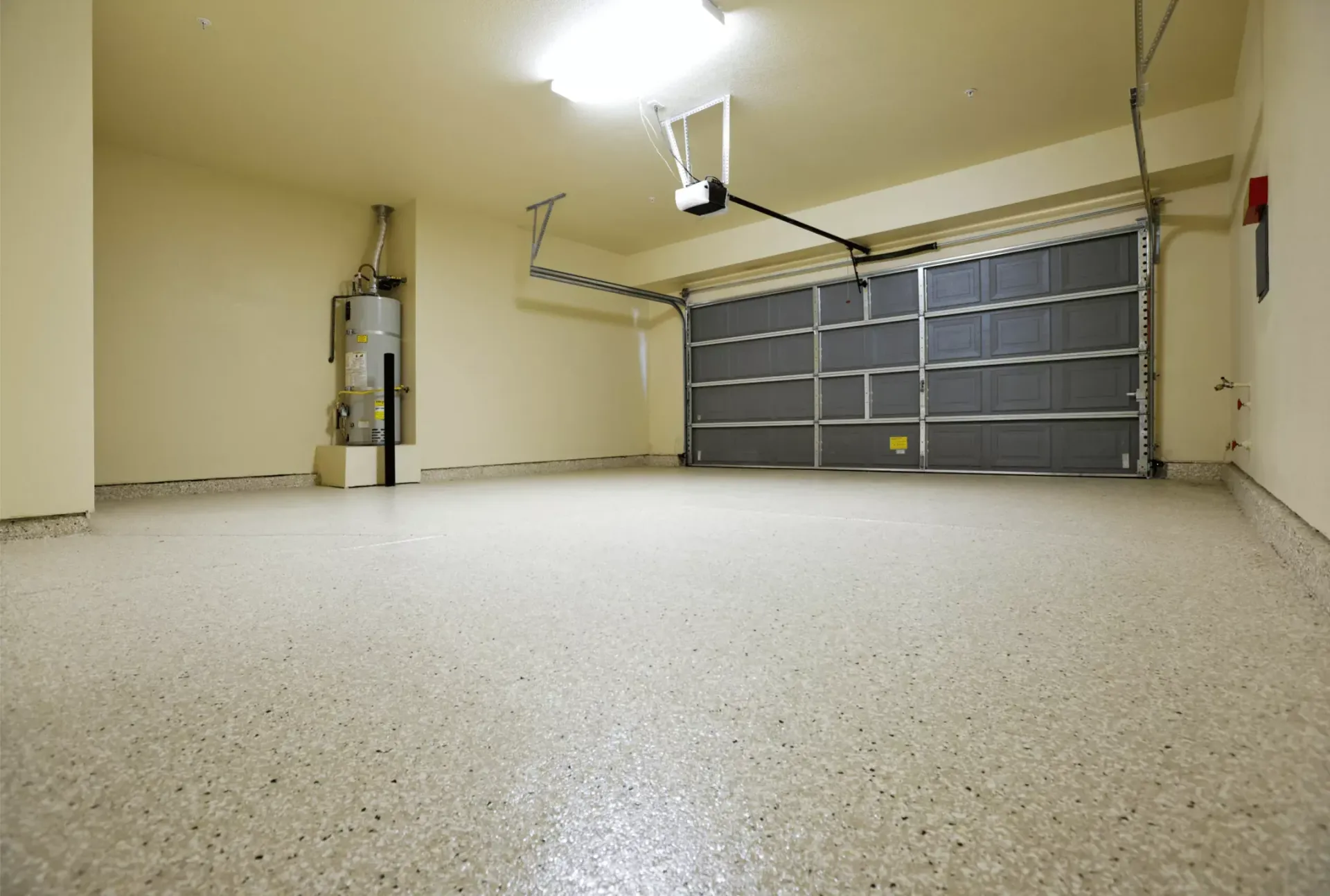 An empty residential garage with beige walls, a speckled epoxy floor, a closed metal door, and a water heater.