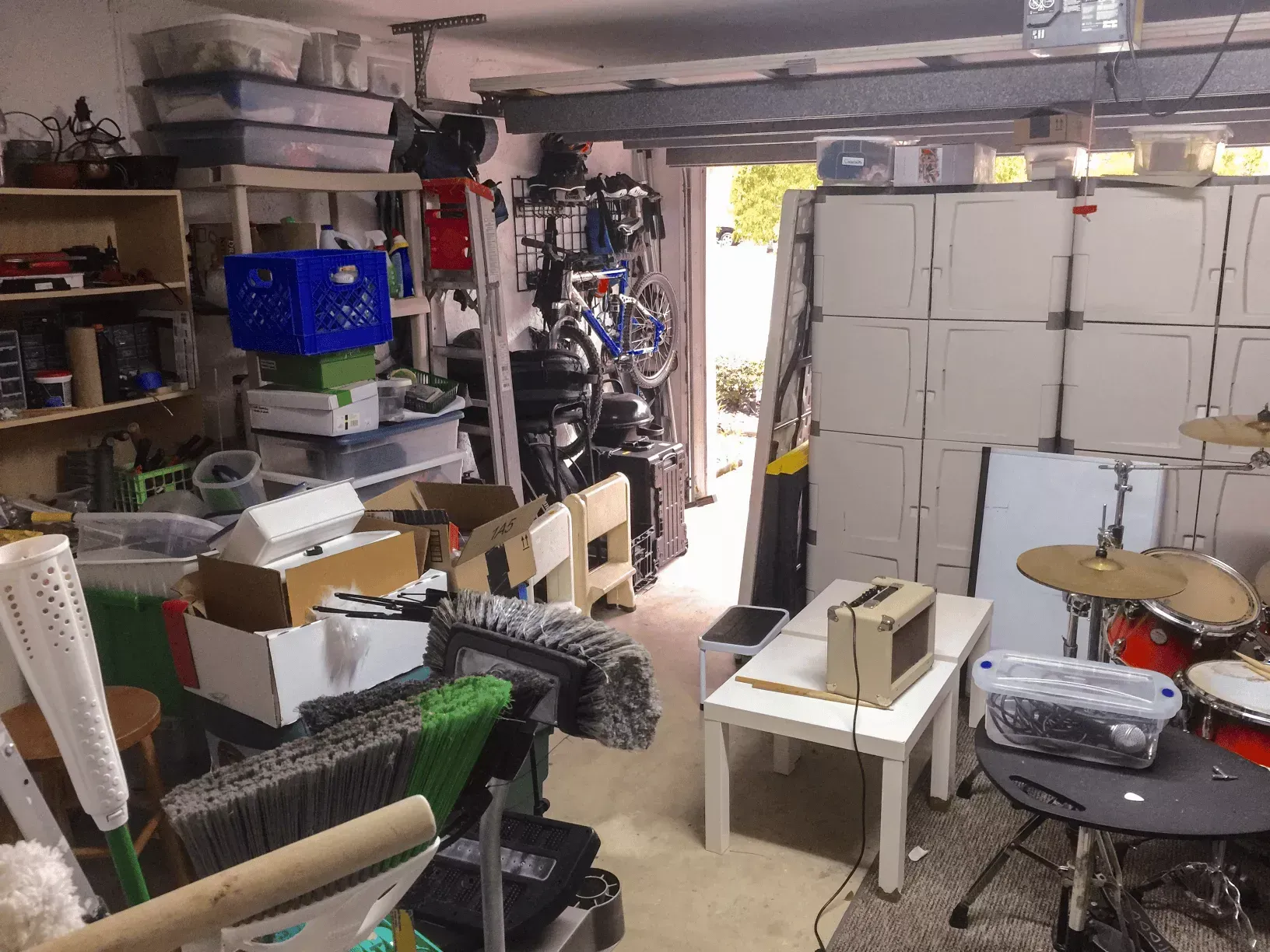 A cluttered garage with storage shelves, a bicycle, white tables, an amplifier, and a drum set near an open doorway.