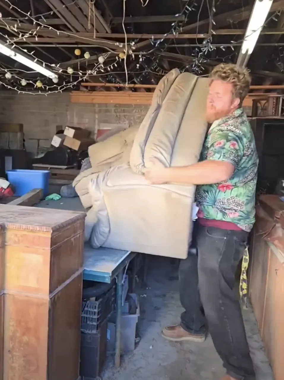 A person in a patterned shirt lifts a large, beige recliner in a cluttered storage garage.