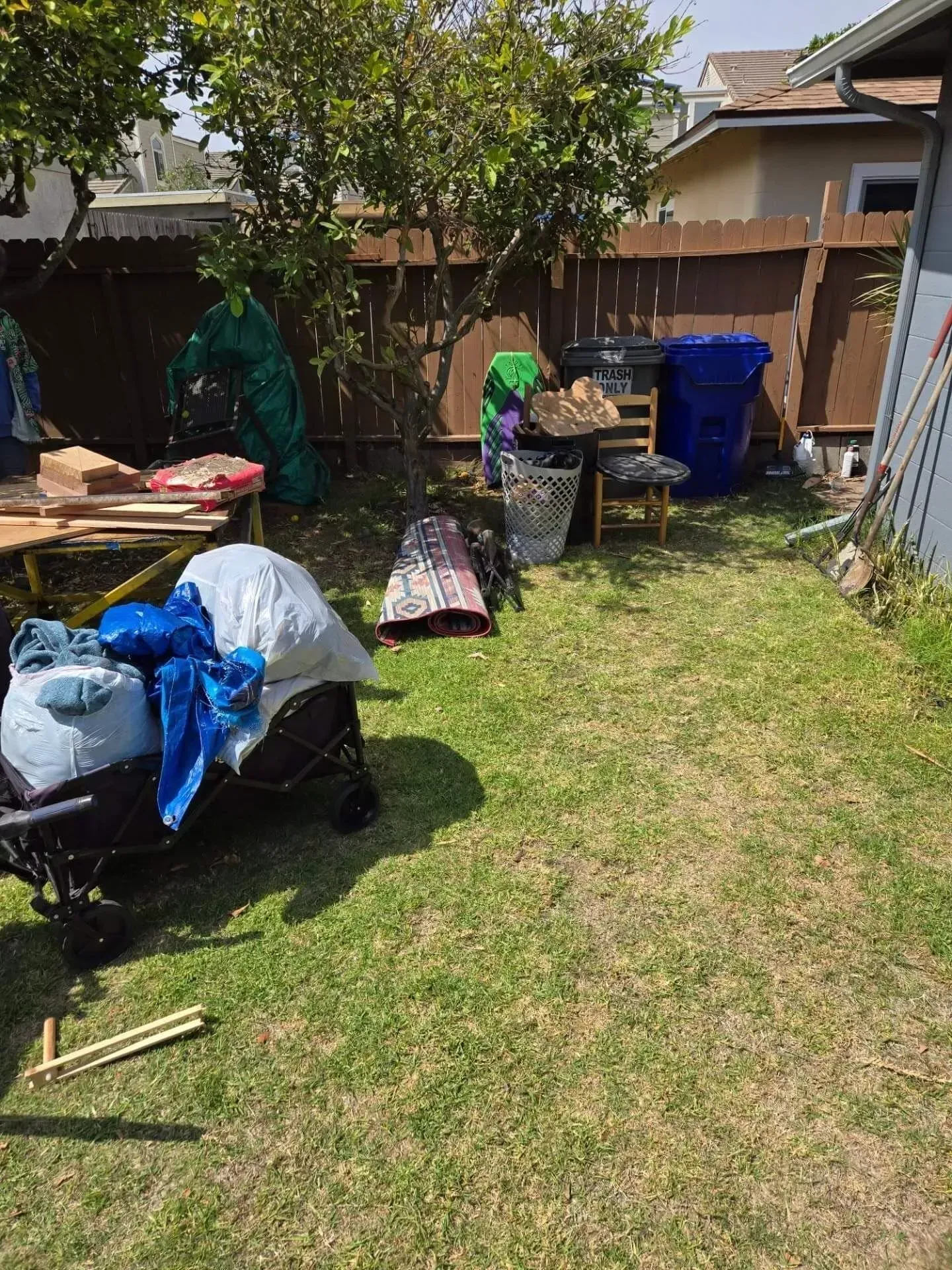 A Ventura beach backyard cluttered with a wagon full of items, a chair, trash bins, and wood scraps near a wooden fence.