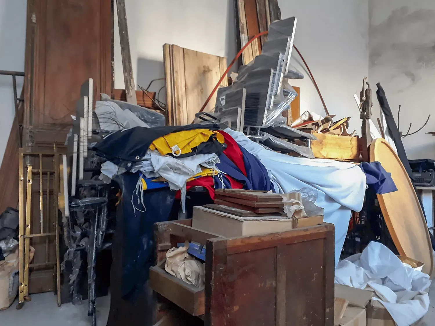 A cluttered storage room filled with miscellaneous wooden boards, metal frames, and draped fabric piles.