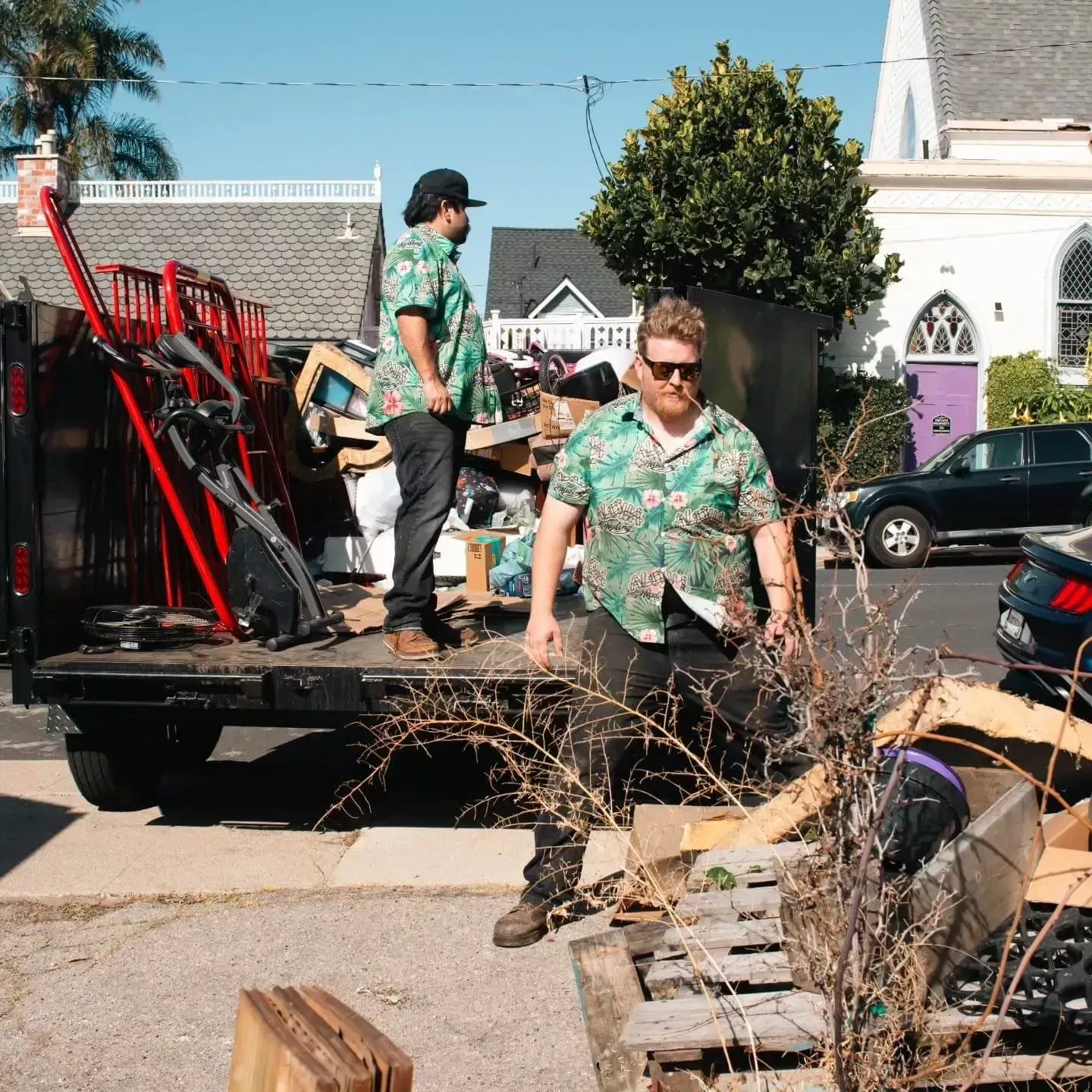 Two people in Hawaiian shirts loading a truck with debris, parked on a street in Ventura, CA. Buildings and a sunny sky in the background.