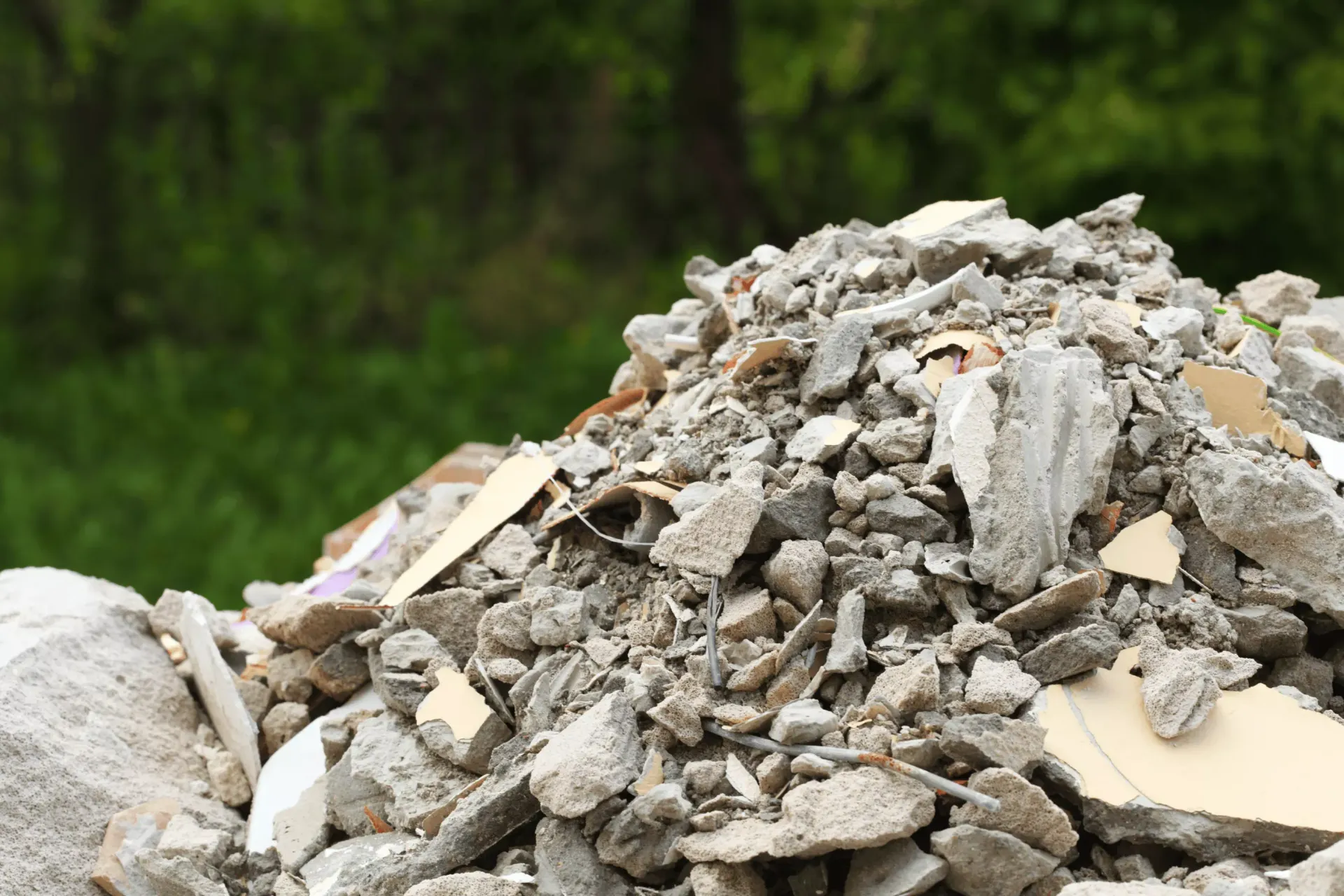 A pile of gray concrete rubble and construction debris sits outdoors in front of a blurred green forest background.