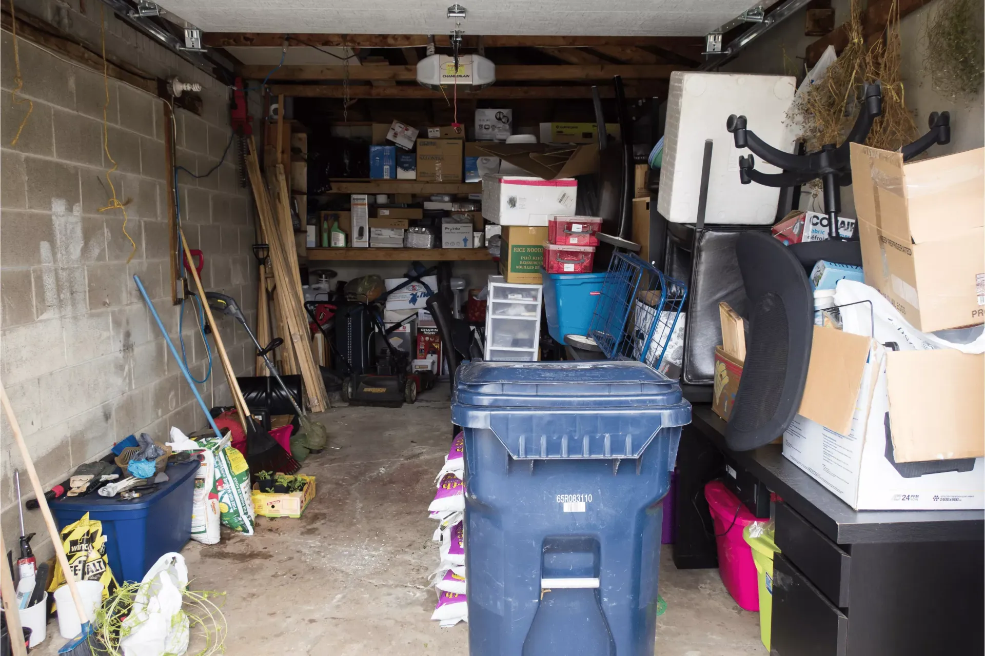 A cluttered residential garage filled with various boxes, tools, a large blue trash bin, and miscellaneous household items.
