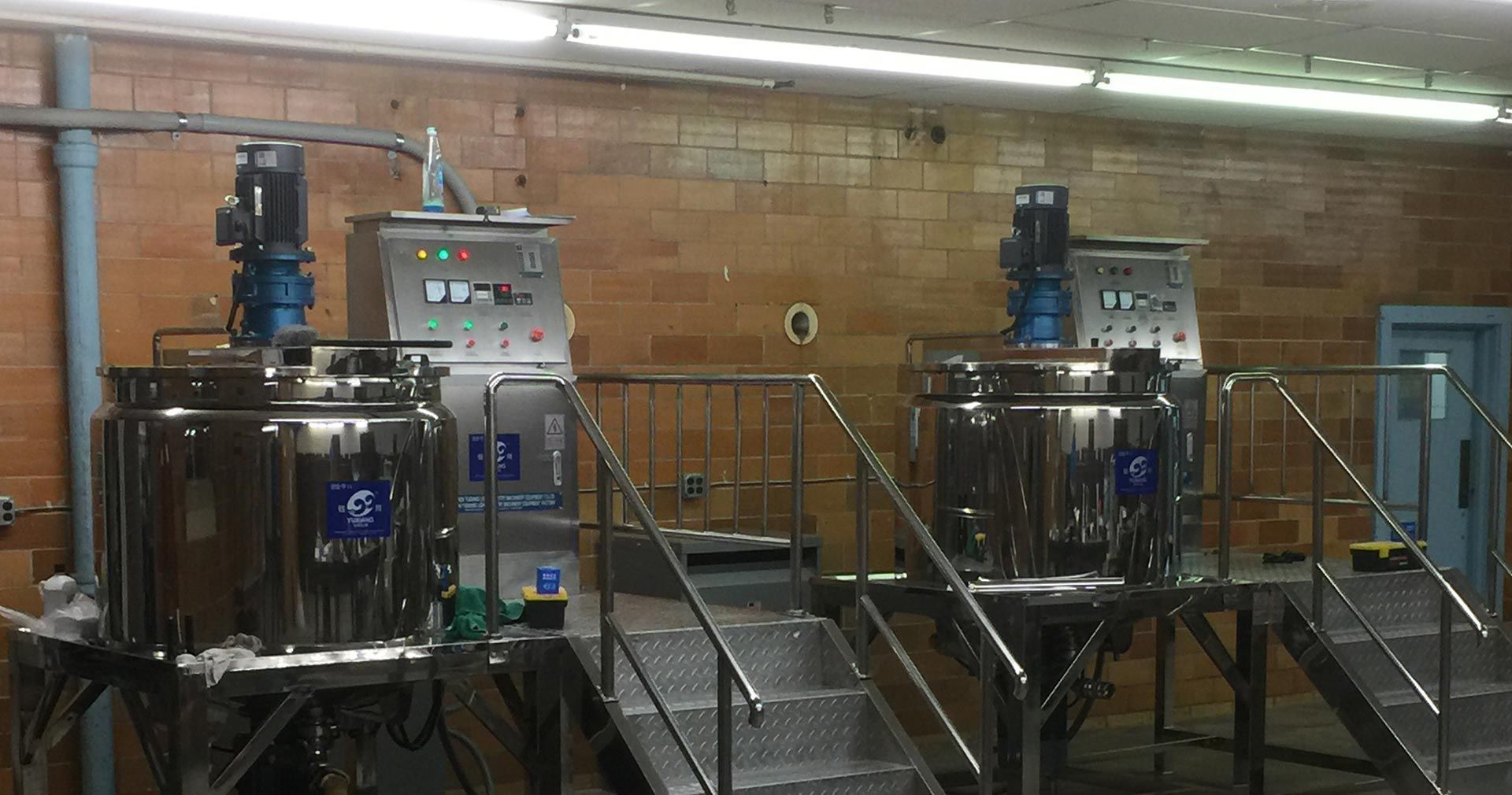 Two stainless steel mixing tanks with control panels on platforms in a factory setting.