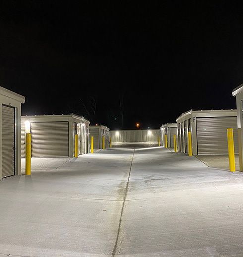 Storage units at night, lit by overhead lights. Gray doors, yellow bollards, dark sky.