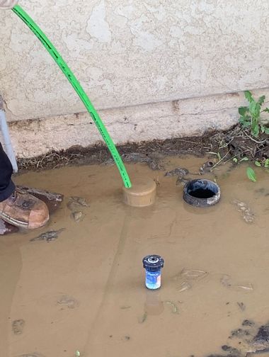 Mud-filled area beside a wall with a green hose, two pipe openings, and a small can on the ground.