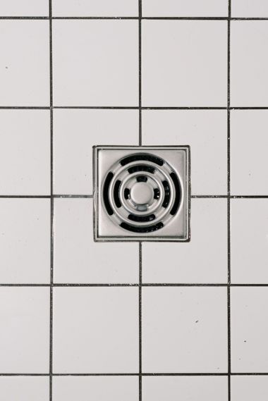 Square metal drain cover in a white tiled shower floor.
