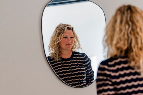 Vrouw kijkt in een elegante spiegel, gekleed in een gestreepte trui, in een witte kamer.