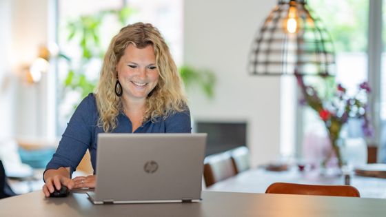 Een vrouw met krullend blond haar glimlacht naar een laptop en gebruikt een muis aan een tafel in een goed verlichte ruimte.