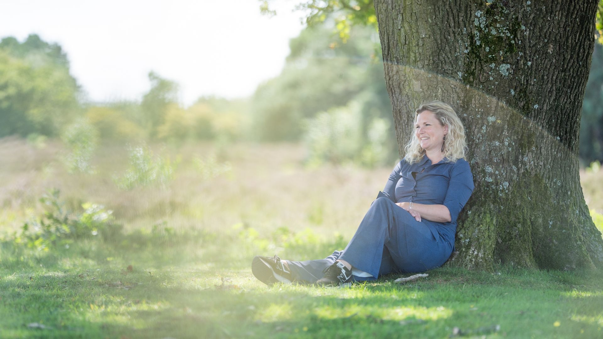Vrouw zit onder een boom, leunend tegen de stam, glimlachend. Buiten, zonnige dag.