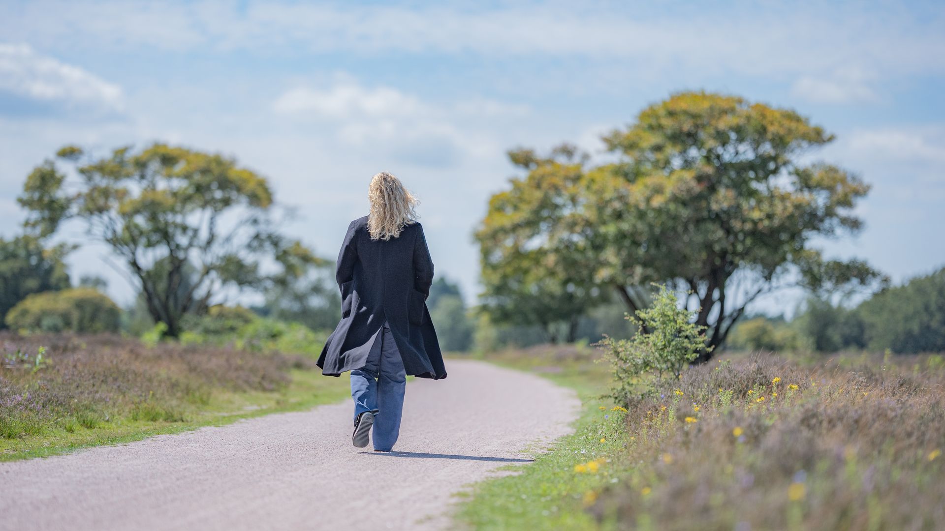 Een vrouw loopt weg over een zandpad, met bomen aan weerszijden, op een zonnige dag.