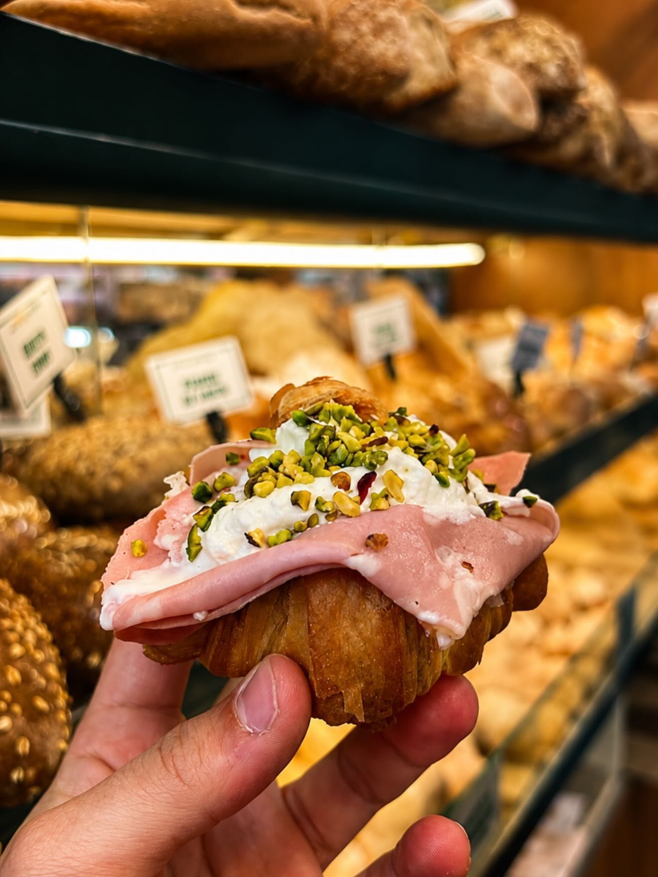 Una mano che regge un pasticcino ricoperto di glassa rosa, pistacchi e noci in una vetrina di pasticceria.