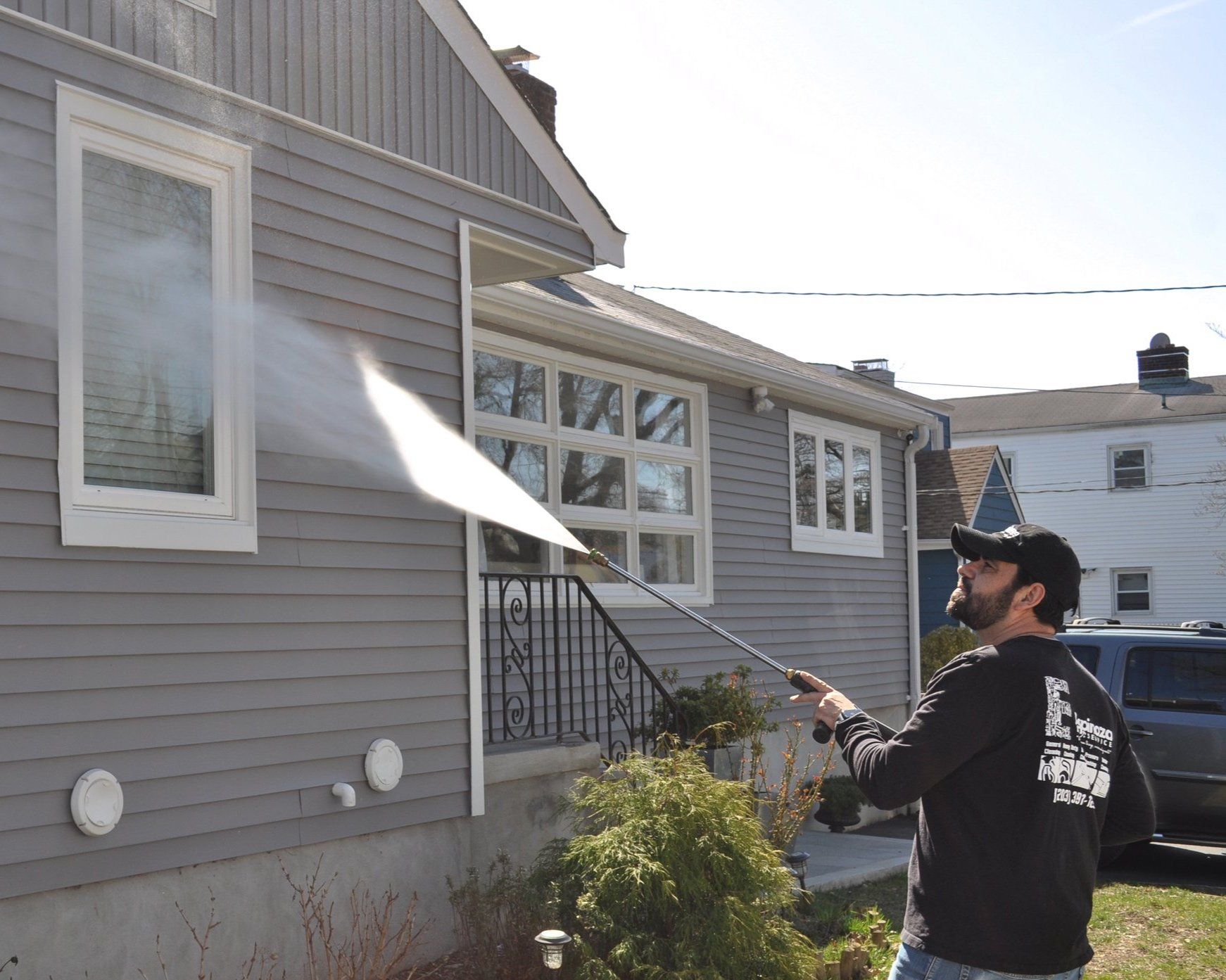 Man pressure-washes the gray house siding near a window with a long spray wand
