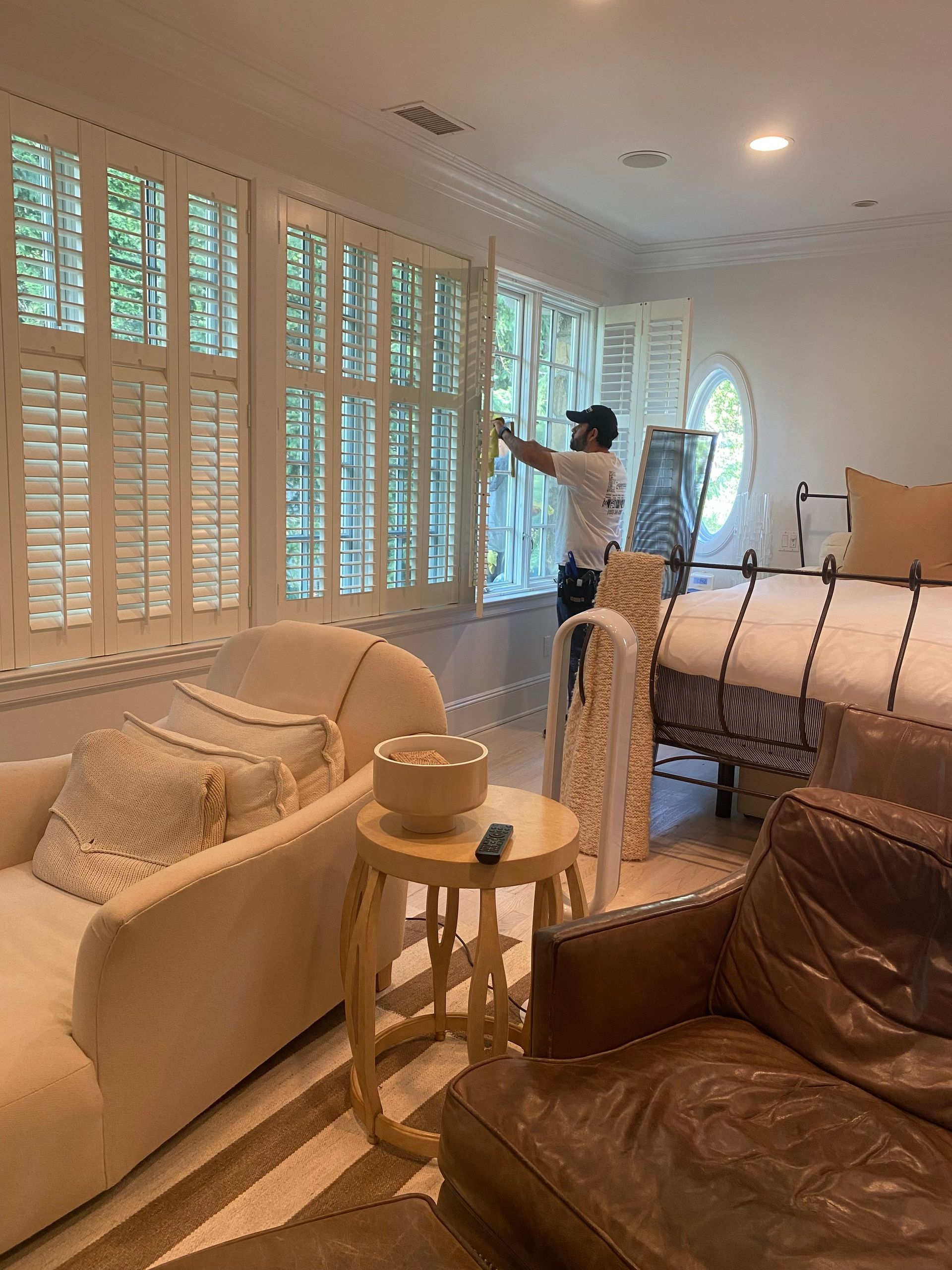 Bright living room with white shutters, cream sofa, brown leather chair, and a person opening the window.