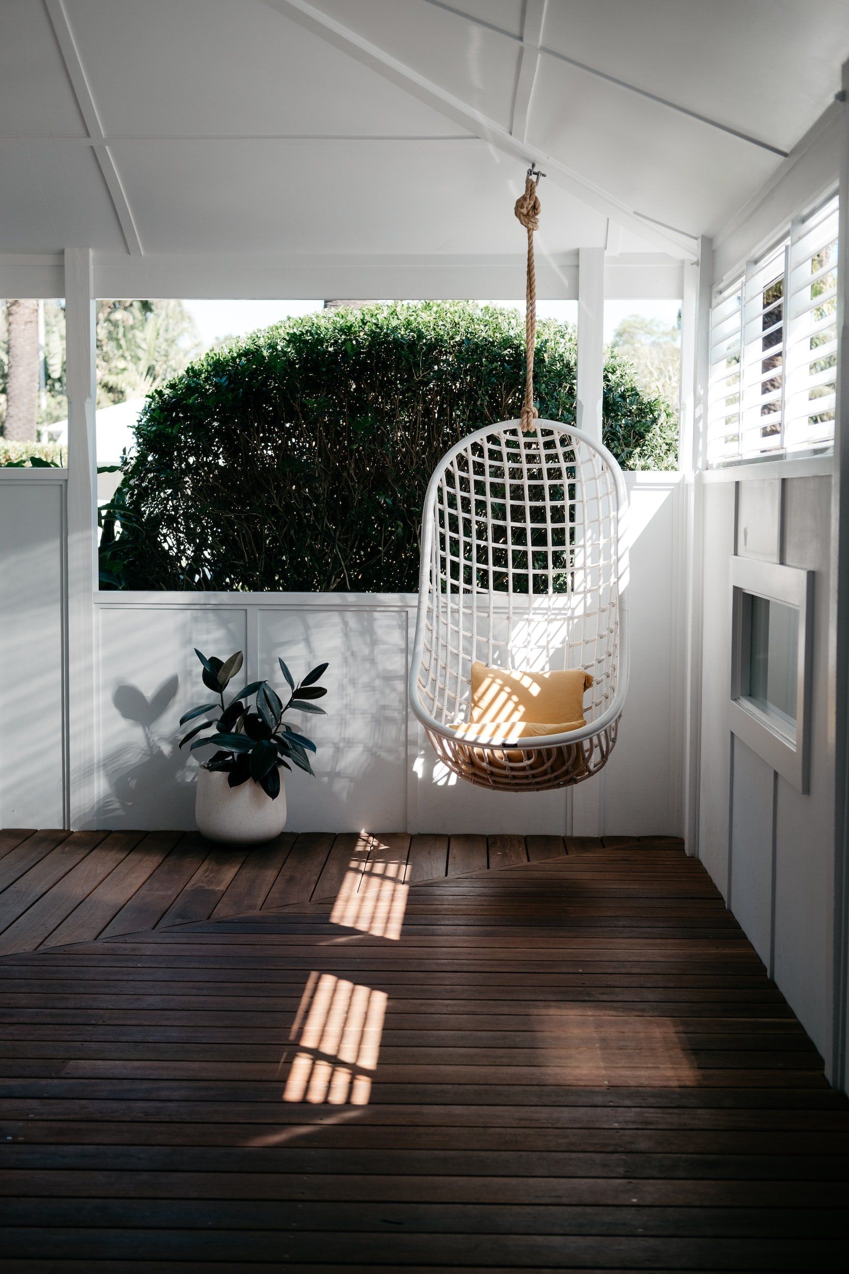 White porch with a hanging chair, potted plant, and sunlight on dark wooden floorboards.