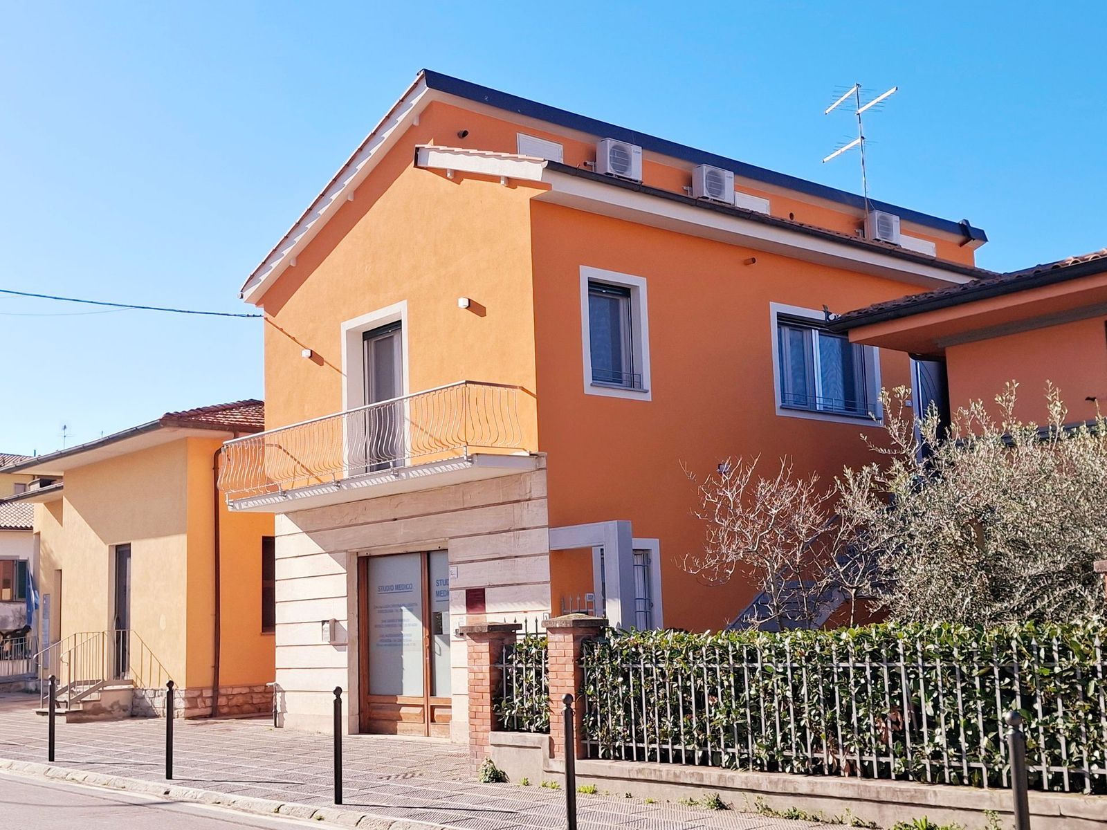 Casa arancione a due piani con balcone, giardino recintato e auto parcheggiata sotto un cielo azzurro e sereno
