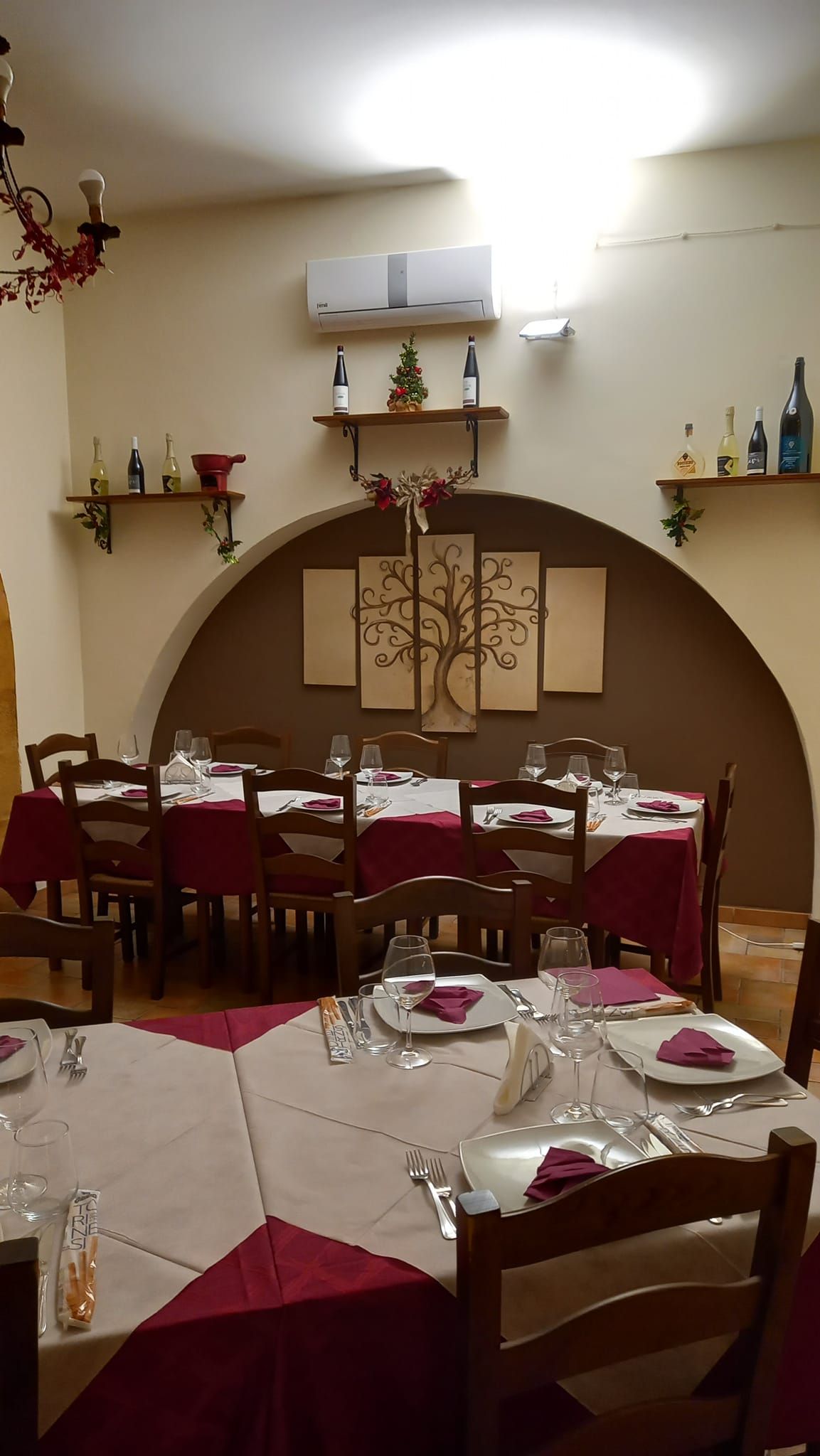 Sala da pranzo del ristorante con tavoli apparecchiati per un pasto; tovaglie rosse, arco, scaffali con decorazioni.