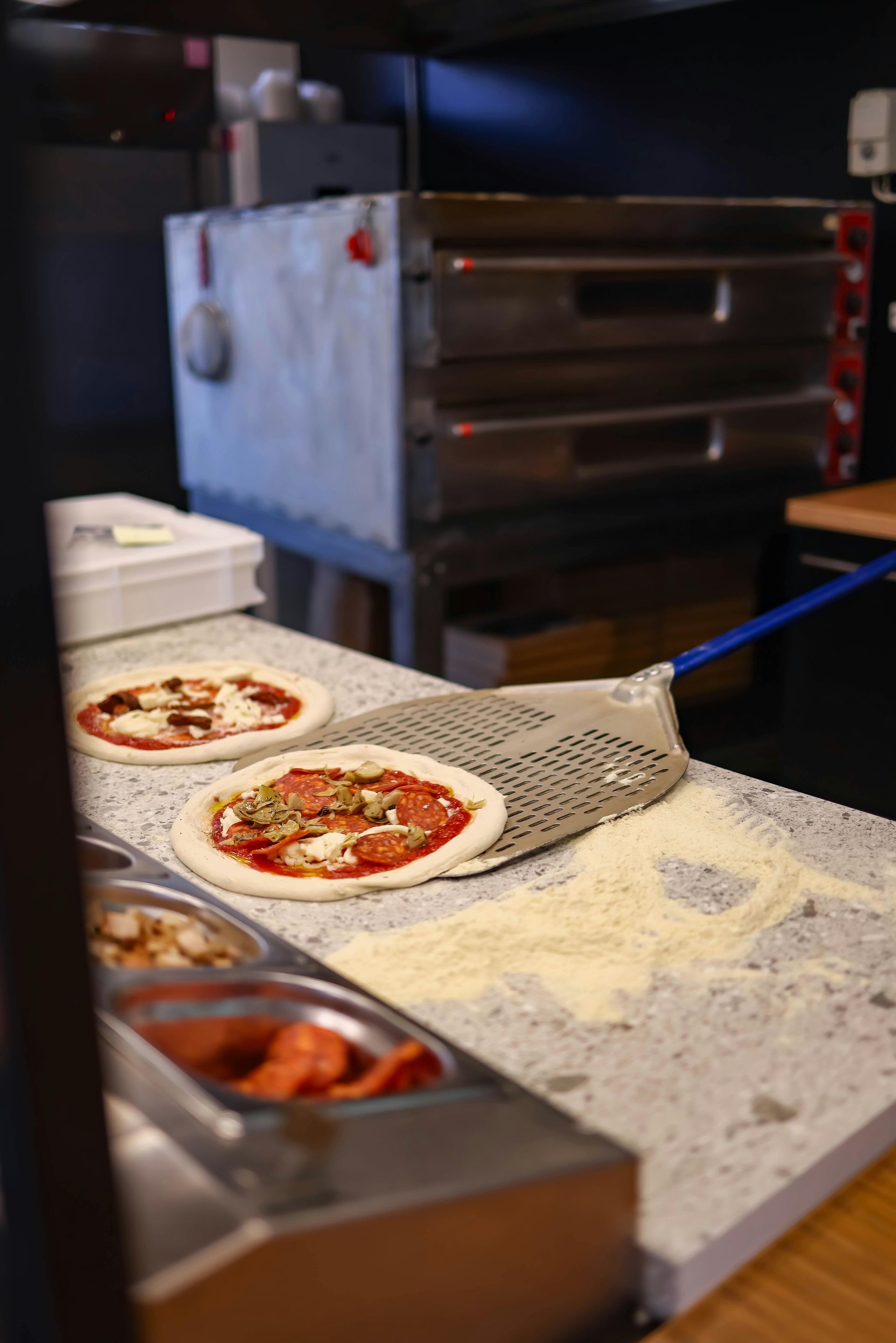 Due pizze su un bancone con condimenti, vicino a un forno e agli ingredienti.