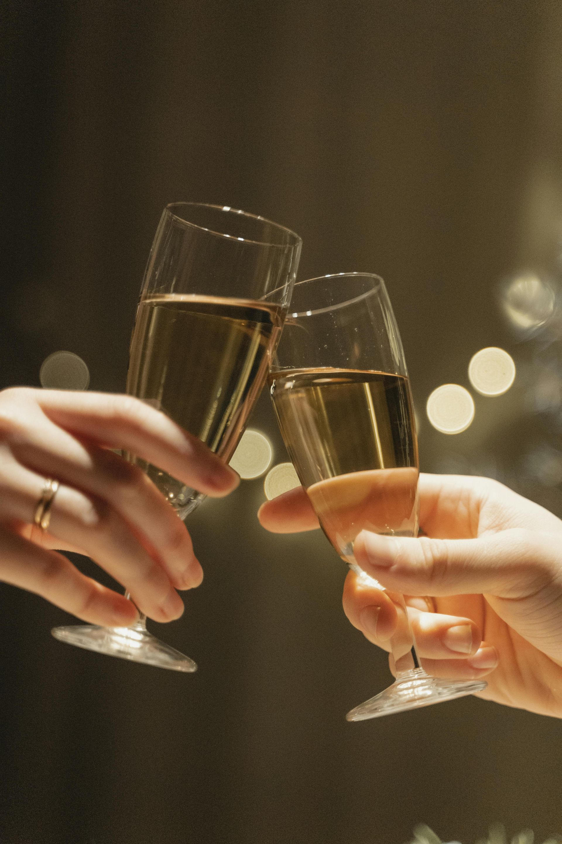 Due mani fanno tintinnare i bicchieri di champagne in un brindisi, mentre le luci delle feste si confondono sullo sfondo.