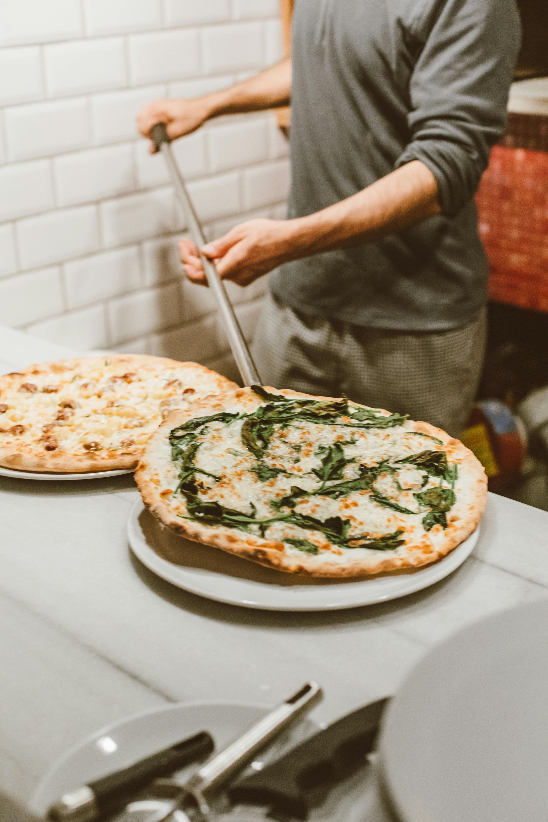 Pizzaiolo con due pizze, una con ingredienti verdi, vicino a un forno a legna.