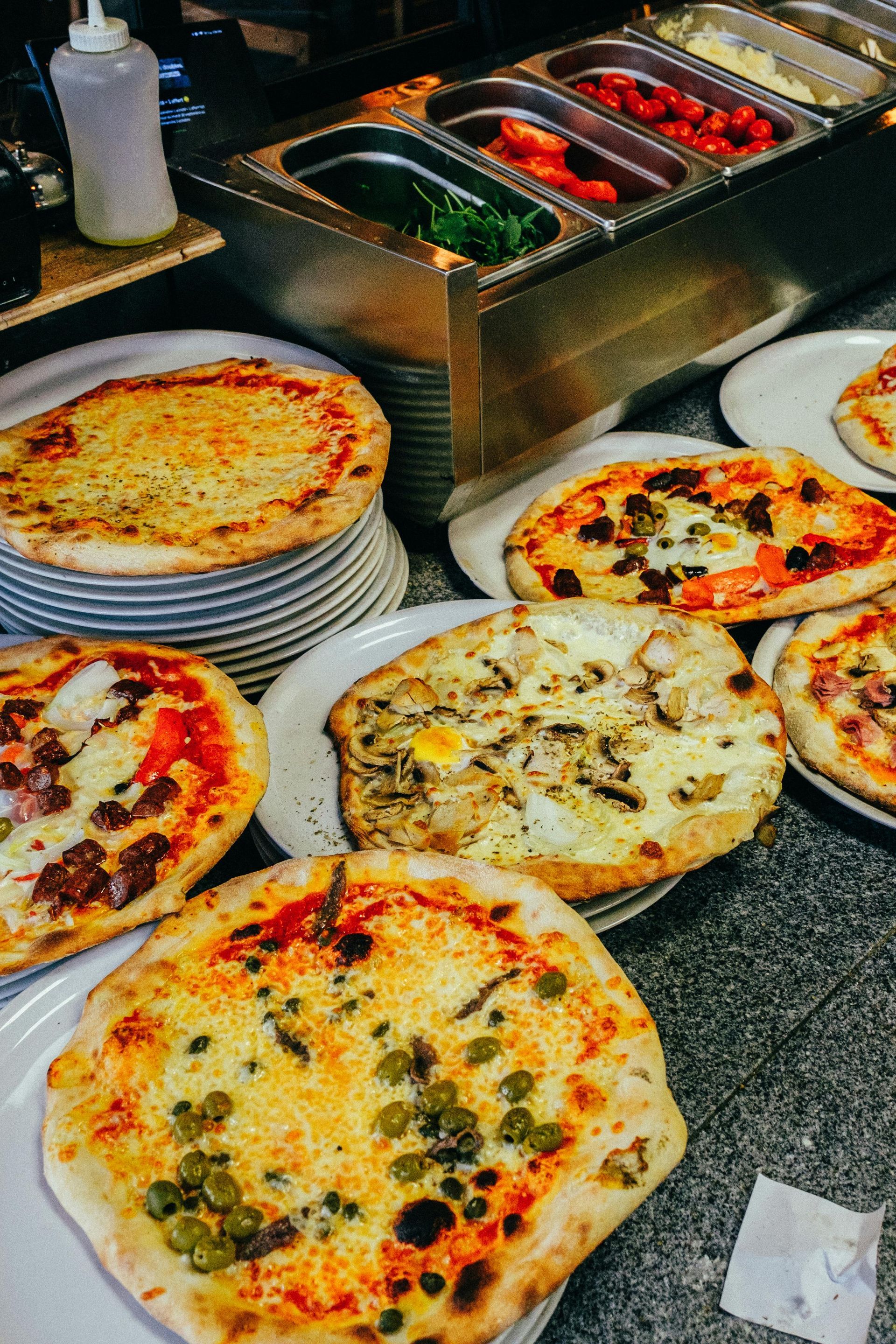 Pizze su piatti bianchi, vicino a un contenitore di condimenti e una pila di pizze.
