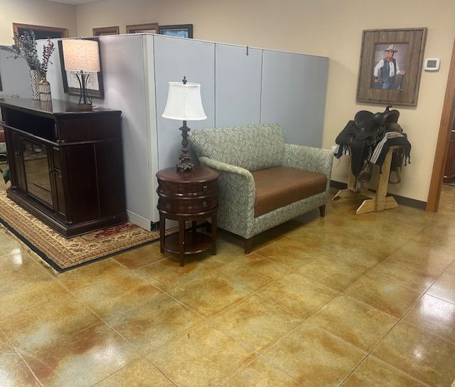 A lobby with tan tile, a gray partition, a patterned loveseat with a brown seat, a side table with a lamp, and a saddle.