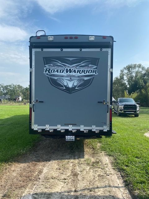 Rear view of a Road Warrior toy hauler trailer with a gray door and logo. A truck is parked nearby.