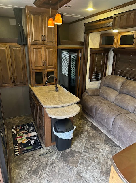 Interior of an RV kitchen featuring a wooden island with a sink, two pendant lights, and a gray sofa nearby.